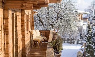 A cozy wooden porch with a chair and a snowy landscape in the background. The trees are covered with snow and the atmosphere is wintry and peaceful.