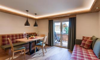 A modern living and dining room with a wooden table and comfortable chairs. Large windows provide access to a balcony and plenty of natural light.