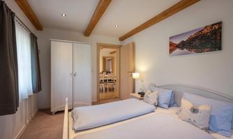 A cozy bedroom with a double bed and soft color tones. In the background, there is an open room with a dining table.
