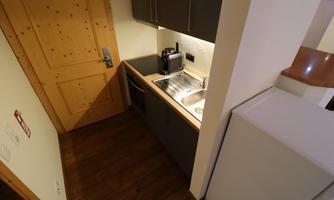 A small, modern kitchen with a wood stove and white walls. In the background, there is a door and a refrigerator.