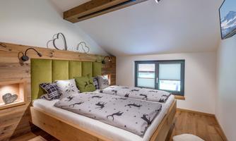 A cozy bedroom with a large bed and a wooden ceiling. The walls are painted in warm tones and there is simple decoration.
