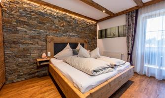 A cozy bedroom with a wooden bed and large pillows. The wall is clad with natural stone and a window provides daylight.