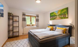 A cozy bedroom with a double bed and a modern shelf. A beautiful landscape painting hangs on the wall and the light is bright and inviting.