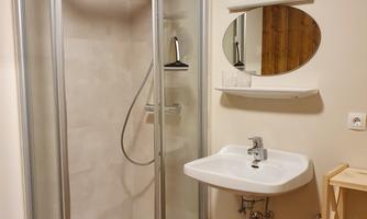 A modern bathroom with a clear shower cubicle and a sink. The walls are bright and the floor has a warm wood tone.