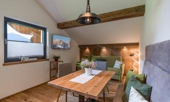 A cozy living area with a wooden table and a green couch. The room has large windows and pleasant lighting.