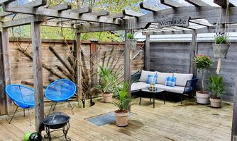 A cozy terrace with wooden posts and an awning. There are comfortable seating arrangements, plants, and a small table.
