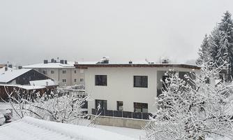 A snowy residential area with white buildings and trees. The sky is gray and the landscape is wintery calm.