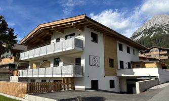 A modern building with wood and stone architecture in the mountains. It has balconies and a beautiful surrounding with green grass.