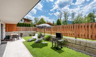 A cozy garden with grass, a sun umbrella, and loungers. In the background, a wooden terrace and a grill can be seen.