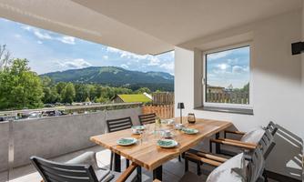 A cozy dining table on a balcony with a view of the mountains. The surroundings are green and inviting under a clear sky.