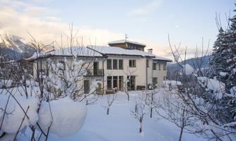 A beautiful house in the snow, surrounded by snow-covered trees. The mountains are visible in the background, and the sky is clear.