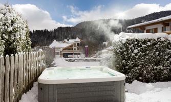 A relaxing hot tub in the snow with steaming water. In the background, snow-capped mountains and characteristic alpine buildings can be seen.