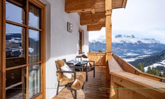 A cozy balcony with a table and chairs, surrounded by wooden paneling. In the background, snow-covered mountains and a clear winter landscape can be seen.