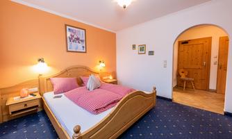 A cozy bedroom with a large bed and checked pillows. The wall is orange, and there is an entrance to a hallway with wooden details.