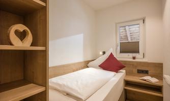 A cozy bedroom with wooden furniture and a window. On the bed lies a pillow with a red border and on the nightstand sits a book.