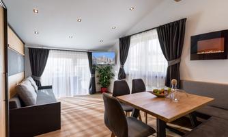 A modern living room with bright windows and elegant curtains. A dining table with chairs and a cozy seating area create an inviting atmosphere.
