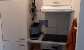 A small, modern kitchen with a stove, a refrigerator, and a range hood. The walls are tiled in light colors and some cooking utensils are hanging.