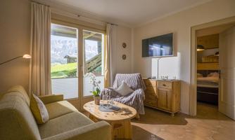 A cozy living room with a green sofa and a wooden table. Large windows provide a view of nature and the mountains.