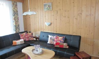 A cozy living room with wooden paneling and two black sofas. In the center is a round table with decorative elements and colorful cushions on the sofas.