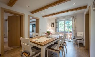 A bright dining room with a large wooden table and white chairs. There are windows that offer a view outside, and the walls are decorated with wooden details.