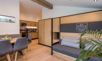 A modern, stylish living room with wooden furniture and a cozy sofa. The room feels bright and inviting, with an open kitchen in the background.