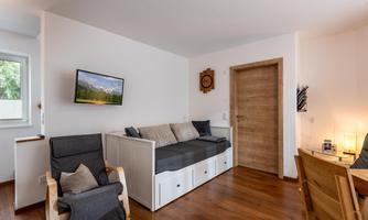 A cozy living space with a sofa bed, a television, and wooden flooring. The walls are bright and inviting, and there is access to another room.