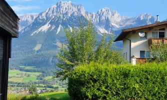 A beautiful view of impressive mountains and green meadows. In the foreground, there are trees and a residential house.