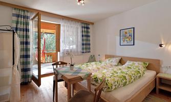 A cozy bedroom with two beds and a small table. The open doors lead to a balcony with a view of the greenery.