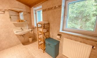 A bright bathroom with white tiles and a window overlooking greenery. There is a sink, a mirror, and a shelf cabinet with towels.