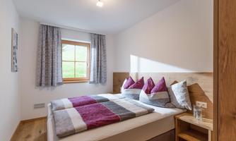 A cozy bedroom with a large bed and colorful pillows. The windows let in soft light and the wooden floor creates a warm atmosphere.