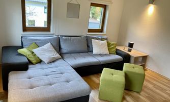 A modern living room with a gray and black sofa and two green stools. There are large windows that let in plenty of daylight.