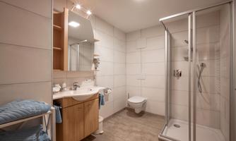 A modern bathroom with a shower, a sink, and a large mirror. There are wooden shelves with blue towels and bright tiles on the walls.