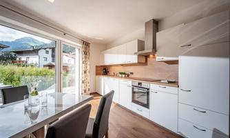 A modern kitchen with white furniture and wooden elements. Large windows offer a view of the surrounding landscape.