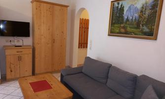 A cozy living area with a gray sofa and a wooden table. A landscape painting hangs on the wall, and a television is installed in the room.