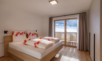 A bright bedroom with modern furniture and large windows. The bed linens are adorned with colorful floral patterns.