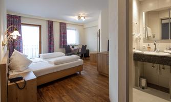 A bright and modern room with two beds and hardwood floors. In the background, a table and chairs, as well as a bathroom, are visible.