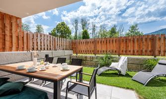 A cozy terrace with a table, chairs, and a beautiful view of the garden. The sun is shining and the sky is partly cloudy.