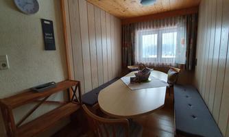 A cozy room with wooden walls and a round table in the middle. There are benches and a large window with a view outside.
