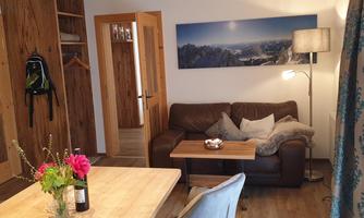 A cozy living room with a leather sofa and a wooden table. A painting of mountains is hanging on the wall, and a bouquet of flowers is on the table.