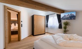 A modern bedroom with wooden details and a large bed. In the background, a television and a view of the mountains are visible.