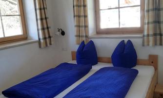 A cozy bedroom with two single beds and blue bedspreads. Large windows provide plenty of daylight.