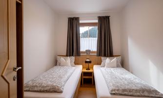 A simple room with two single beds, a nightstand, and a window. The walls are light and the curtains are dark.