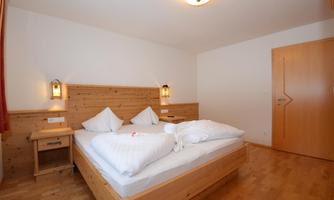 A cozy bedroom with a wooden double bed. The walls are painted light and there is soft lighting.