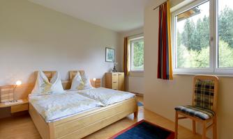 A cozy bedroom with a double bed and two nightstands. The room has large windows overlooking greenery and a colorful curtain.