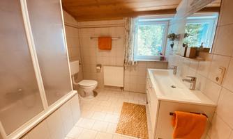 A bright bathroom with white tiles and a wooden ceiling. It features a shower, toilet, sink, and a window with a view of the outdoors.
