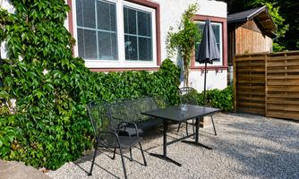 A cozy outdoor area with a table and two chairs, surrounded by green plants. In the background, there is a window and a wooden slatted wall.