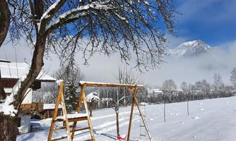 A snow-covered garden with a swing and a tree. In the background, mountains and a bright blue sky can be seen.