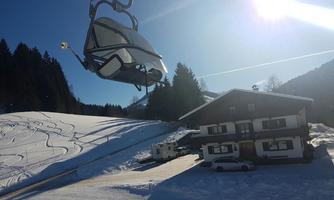 A sunny winter landscape with a ski lift and snow-covered hills. In the foreground stands a cozy mountain house.