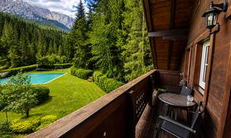 A beautiful view from a balcony overlooking a green meadow with a pool. In the background, majestic mountains and dense trees can be seen.