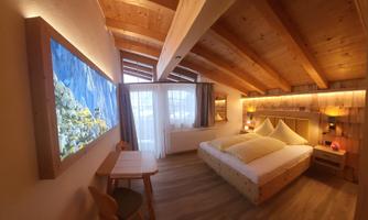 A cozy bedroom with a wooden ceiling and warm light. There is a large bed, a table, and a window with a view.